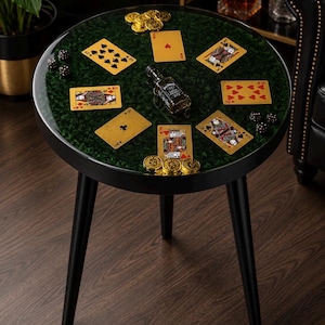 Könnte beinhalten: Ein runder schwarzer Beistelltisch mit drei konischen Beinen und einer dunkelgrün marmorierten Tischplatte. Die Oberfläche ist mit goldenen Spielkarten, mehreren Goldmünzen, einer kleinen Glasflasche Jack Daniel's Whiskey und schwarzen Würfeln dekoriert.