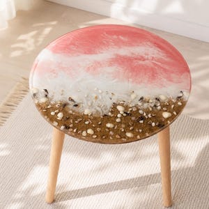 Pode incluir: Uma mesa lateral redonda com três pernas de madeira clara e um tampo circular com um design em resina. A superfície apresenta um padrão de redemoinho rosa e branco que lembra ondas encontrando uma costa arenosa com pequenas conchas.