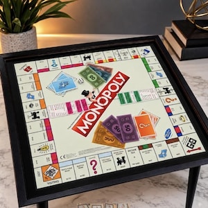 May include: A square Monopoly board game table with a black frame and tapered legs. The board features the classic Monopoly logo, colorful property spaces, Chance and Community Chest cards, paper money in various denominations, and a die.