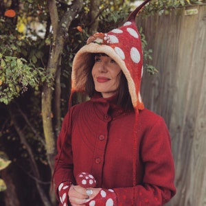 May include: A red mushroom-themed hat with white polka dots, a matching mushroom-shaped accessory, and red gloves with white polka dots. The hat has a pointed top and a beige interior. The person is wearing a red coat.