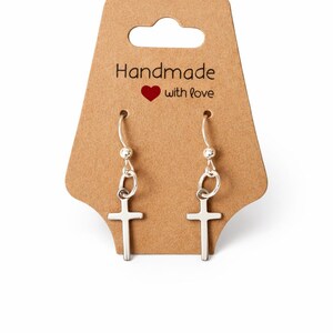 May include: A pair of silver-colored cross earrings hanging from a silver hook. The earrings are displayed on a brown card with the text "Handmade with love" and a red heart.