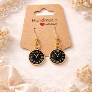 May include: A pair of gold-toned clock face earrings with black faces and Roman numeral hour markers. The earrings are attached to a brown card that reads "Handmade with love". The background includes lace and decorative elements.