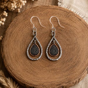 May include: A pair of dangle earrings featuring a silver-toned hammered teardrop frame surrounding a smaller, dark, intricate filigree teardrop charm with a copper-toned edge. The earrings are attached to clear plastic fishhook-style ear wires.