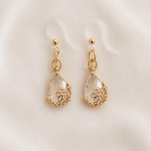 May include: A pair of gold-toned earrings with clear, teardrop-shaped pendants. Each pendant features a small gold flower and is connected to a gold chain and a clear ear hook. The earrings are displayed on a white background.