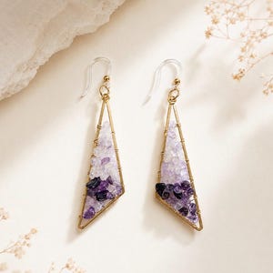 May include: A pair of dangle earrings featuring gold-toned triangular wire frames filled with small, tumbled amethyst gemstone chips in varying shades of light and dark purple. The earrings are attached to clear plastic hook-style ear wires.