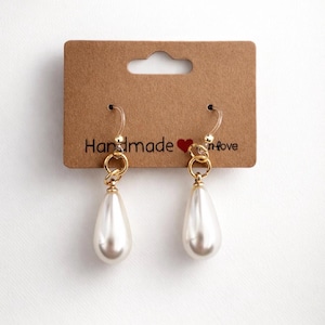 May include: A pair of handmade pearl drop earrings with gold-colored accents. The earrings are displayed on a brown card with the words "Handmade" and "love" printed on it. The earrings have a teardrop shape.