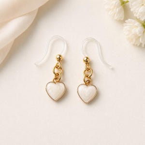 May include: A pair of gold-colored earrings with white heart-shaped pendants. The earrings have clear plastic ear wires and are displayed on a cream-colored surface. The heart pendants have a gold outline and a swirled white enamel filling.