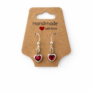 May include: A pair of heart-shaped earrings with a vibrant red center, set in a silver-toned metal. The earrings are displayed on a brown card with the text "Handmade with love".