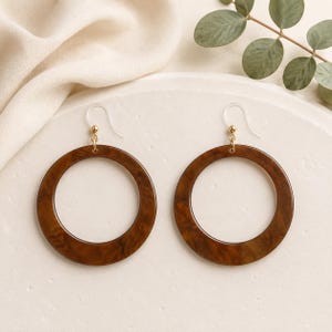 May include: A pair of circular drop earrings featuring a dark brown tortoiseshell pattern. The earrings are suspended from clear plastic fishhook wires with small gold-toned metal connectors, creating a simple and balanced design for these round accessories.