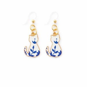May include: A pair of cat-shaped earrings. Each earring features a white cat silhouette with blue floral accents and a gold-colored metal loop. The earrings hang from clear, plastic ear wires. The design is simple and elegant.