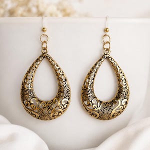 May include: A pair of gold-toned teardrop-shaped earrings featuring an intricate openwork filigree pattern. The earrings are suspended from small circular jump rings and clear plastic ear hooks, showcasing a detailed metal scrollwork design.