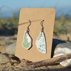 May include: Handmade earrings with irregular white ceramic pieces, adorned with green and blue patterns. The earrings, suspended from silver hooks, are displayed on a brown card. The background is a sandy beach, with shells and pebbles.