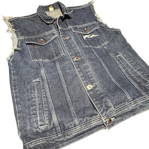 May include: A dark blue denim vest with a classic collar and button-up front. Features two chest pockets and a small embroidered design on the right pocket. The vest has distressed edges and a vintage look.