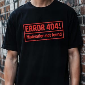 Può includere: T-shirt nero con grafica rettangolare rossa con la scritta "ERROR 404! Motivation not found." La maglietta ha maniche corte e girocollo. L'immagine è un primo piano del petto.
