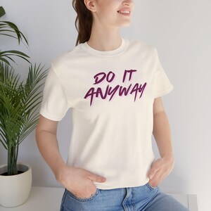 Könnte beinhalten: Ein cremefarbenes Kurzarm-T-Shirt mit dem Text "DO IT ANYWAY", der in einer dunkelvioletten Schriftart mit einem hellvioletten Schatteneffekt gedruckt ist. Das T-Shirt hat einen Rundhalsausschnitt und wird mit einer blauen Jeans kombiniert.