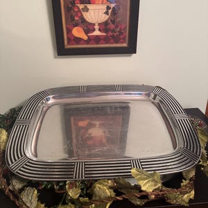 Puede incluir: Una bandeja rectangular plateada con un borde a rayas negras y plateadas. Encima de la bandeja hay un cuadro enmarcado de un cuenco de fruta. El cuadro tiene un marco negro y está colgado en una pared verde claro.