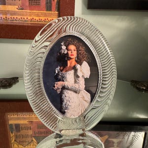 May include: An oval-shaped clear glass picture frame featuring a textured, wavy, rippled pattern throughout the border. The frame holds a photograph of a person with voluminous hair wearing a white, textured, long-sleeved garment.