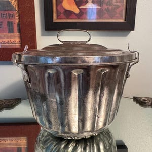 May include: A vintage, silver-toned metal mold with a lid and handle. The mold has a fluted design and is reflected in the surface below. The piece is likely used for baking or food preparation.
