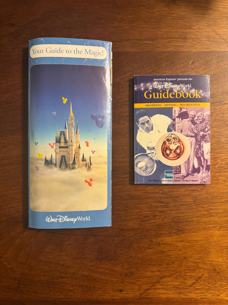 K&ouml;nnte beinhalten: Zwei Vintage-Reisef&uuml;hrer von Walt Disney World. Einer ist eine blaue Brosch&uuml;re mit einer Schlossillustration und dem Text "Your Guide to the Magic!". Der andere ist ein Reisef&uuml;hrer mit dem Text "Guidebook" und "American Express presents the Walt Disney World".
