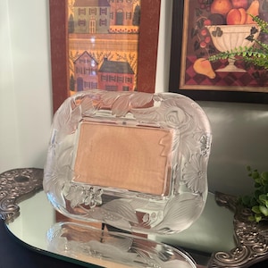 May include: A rectangular glass picture frame featuring a frosted, etched floral design along its border. The frame has a clear, smooth center area for a photograph and is supported by a stand, reflecting on a mirrored surface below.