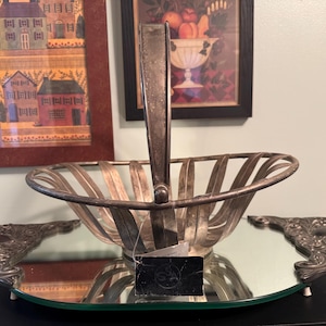 May include: Silver-toned metal basket with a handle, set on a mirrored tray with ornate edges. The basket has a slatted design. Decorative artwork hangs in the background.