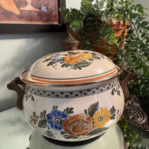 Könnte beinhalten: Cremefarbener Keramik-Bräter mit Deckel, verziert mit bunten Blumenmustern in Blau, Orange und Gelb. Der Topf hat braune Griffe und einen passenden Knauf. Ein dekorativer Küchenartikel.