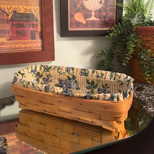 Puede incluir: Una cesta rectangular de madera tejida con un acabado marrón claro y un forro de tela. El forro está decorado con un patrón de rosas azules y hojas verdes sobre un fondo de finas rayas verticales amarillas y blancas.