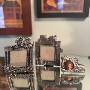 May include: Three small, silver-toned picture frames. One frame has a dog and cat design, another has a floral design, and the third is shaped like a baby boot. The frames are on a reflective surface.