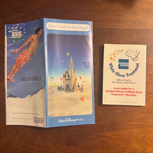K&ouml;nnte beinhalten: Zwei Brosch&uuml;ren und ein B&uuml;chlein auf einer Holzoberfl&auml;che. Eine Brosch&uuml;re zeigt einen Mann beim Skifahren mit dem Text "SKI DISNEY" und "Blizzard Beach water park". Die andere Brosch&uuml;re sagt "Your Guide to the Magic!" und zeigt ein Schloss. Das B&uuml;chlein sagt "White Glove Treatment".