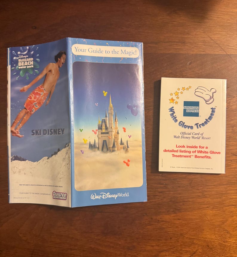 K&ouml;nnte beinhalten: Zwei Brosch&uuml;ren und ein B&uuml;chlein auf einer Holzoberfl&auml;che. Eine Brosch&uuml;re zeigt einen Mann beim Skifahren mit dem Text "SKI DISNEY" und "Blizzard Beach water park". Die andere Brosch&uuml;re sagt "Your Guide to the Magic!" und zeigt ein Schloss. Das B&uuml;chlein sagt "White Glove Treatment".