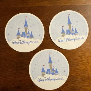 K&ouml;nnte beinhalten: Drei runde Untersetzer mit hellblauem Hintergrund, einem Schlossdesign und dem Text "Walt Disney World". Das Schloss hat blaue T&uuml;rme, einen beigen K&ouml;rper und rote Fahnen. Die Untersetzer sind wei&szlig; mit einem hellblauen Rand.