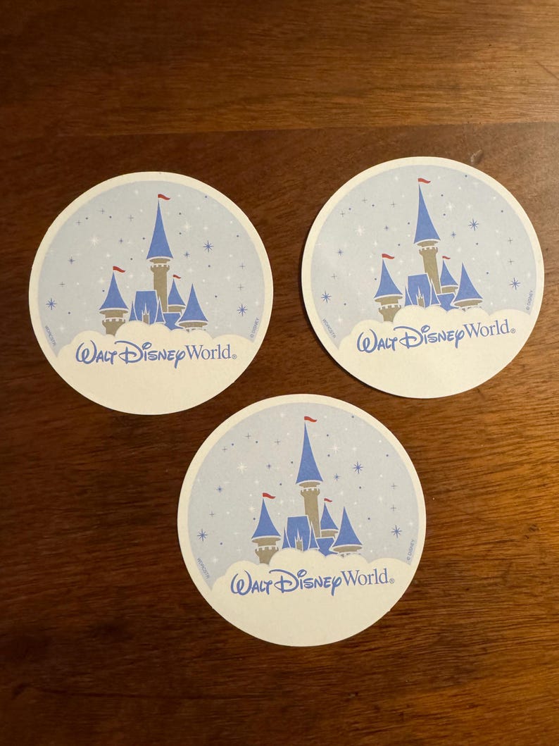 K&ouml;nnte beinhalten: Drei runde Untersetzer mit hellblauem Hintergrund, einem Schlossdesign und dem Text "Walt Disney World". Das Schloss hat blaue T&uuml;rme, einen beigen K&ouml;rper und rote Fahnen. Die Untersetzer sind wei&szlig; mit einem hellblauen Rand.