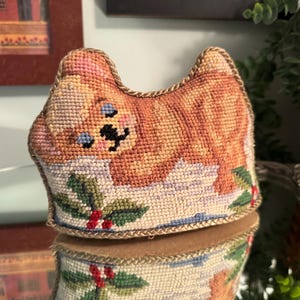 May include: A cat-shaped needlepoint pincushion featuring a tan cat with blue eyes lying down on a snowy surface decorated with green holly leaves and red berries. The edges are finished with a braided gold trim and a brown felt base.