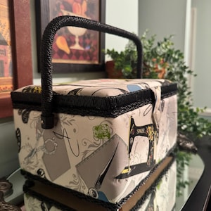 May include: A vintage sewing box with a black handle and trim. The box is covered in fabric with a pattern of sewing tools, including scissors and a sewing machine. The word "SINGER" is visible on the side.