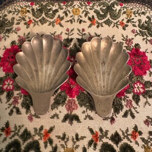 May include: Two silver-colored, shell-shaped dishes. The dishes have a fan-like design with etched details. They rest on a floral patterned fabric with red, green, and yellow flowers.