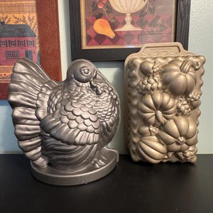 May include: Two decorative metal baking pans. One is shaped like a turkey, the other is rectangular with a harvest theme, featuring pumpkins, gourds, and corn. The turkey pan is silver, and the harvest pan is gold.