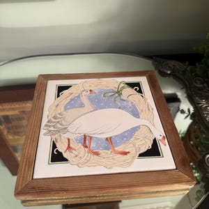 May include: A square ceramic tile framed in light brown wood featuring an illustration of two white geese with orange beaks and feet. The geese are positioned inside a tan woven wreath against a blue background with white polka dots.