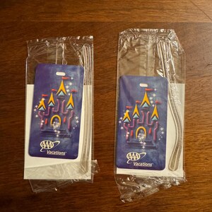 K&ouml;nnte beinhalten: Zwei AAA Vacations Gep&auml;ckanh&auml;nger in Kunststoffverpackung. Jeder Anh&auml;nger zeigt ein Schlossdesign mit gelben, orangefarbenen und blauen Akzenten auf dunklem blauem Hintergrund. Die Anh&auml;nger sind rechteckig mit einem Loch oben.