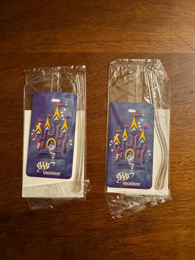 K&ouml;nnte beinhalten: Zwei AAA Vacations Gep&auml;ckanh&auml;nger in Kunststoffverpackung. Jeder Anh&auml;nger zeigt ein Schlossdesign mit gelben, orangefarbenen und blauen Akzenten auf dunklem blauem Hintergrund. Die Anh&auml;nger sind rechteckig mit einem Loch oben.
