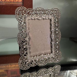 May include: A rectangular picture frame with a metallic silver finish and an intricate, openwork filigree border featuring scrolling patterns and floral motifs. The frame has a plain, light-colored backing insert and a reflective surface.