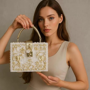 Könnte beinhalten: Eine weiße und goldene Box-Handtasche mit goldenem Griff und Verschluss. Die Handtasche ist mit goldenen floralen Designs, kleinen klaren Edelsteinen und zwei goldenen Medaillons verziert. Die Handtasche wird von einer Person gehalten.