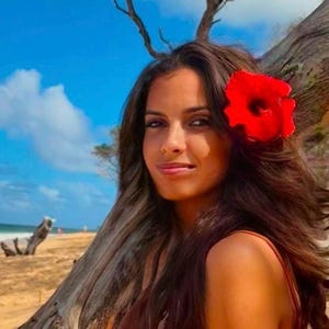 May include: A woman with long brown hair and a red flower tucked behind her ear. She is on a beach with a blue sky and a tree in the background. The woman is wearing a red top.