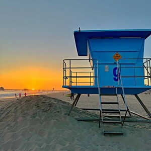 Könnte beinhalten: Ein blauer Rettungsturm steht am Strand bei Sonnenuntergang. Der Himmel ist ein Farbverlauf von Orange und Blau. Eine Leiter und Geländer sind am Turm befestigt. Menschen und ein Pier sind in der Ferne zu sehen.