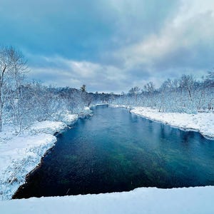 Könnte beinhalten: Eine ruhige Winterlandschaft mit einem Fluss, der durch einen schneebedeckten Wald fließt. Das Wasser ist ein tiefes Blaugrün, das den bewölkten Himmel widerspiegelt. Kahle Bäume säumen die Ufer, bedeckt mit Schnee, was eine ruhige, kalte Szene schafft.