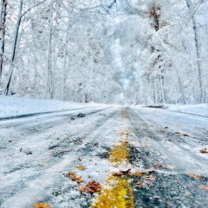 Könnte beinhalten: Eine schneebedeckte Straße mit gelber Mittellinie führt in einen Wald aus schneebedeckten Bäumen. Die Fahrbahn ist teilweise mit Eis und abgefallenen Blättern bedeckt und schafft eine Winterszene.