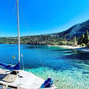 Könnte beinhalten: Ein weißes Segelboot mit blauem Segel liegt in klarem, türkisfarbenem Wasser vor Anker. Das Boot befindet sich in der Nähe eines Holzstegs, mit einem Hintergrund aus bewaldeten Bergen und einem strahlend blauen Himmel. Die Szene vermittelt Ruhe und Erholung im Freien.