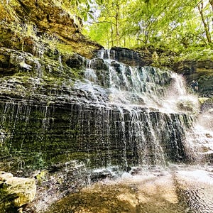K&ouml;nnte beinhalten: Ein Wasserfall st&uuml;rzt &uuml;ber geschichtete Felsformationen und bildet eine nat&uuml;rliche Kaskade. Das Wasser ist klar und reflektiert das Sonnenlicht. &Uuml;ppiges gr&uuml;nes Laub umrahmt die Spitze des Wasserfalls. Eine Person steht in der N&auml;he des Fu&szlig;es.