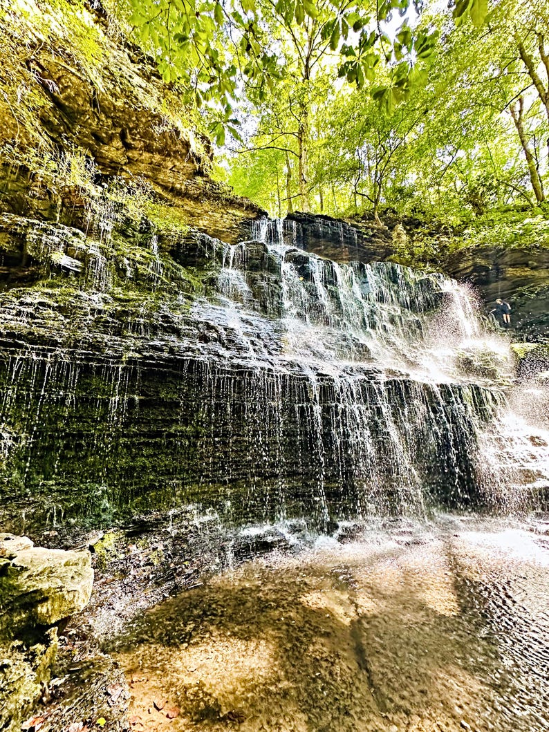 K&ouml;nnte beinhalten: Ein Wasserfall st&uuml;rzt &uuml;ber geschichtete Felsformationen und bildet eine nat&uuml;rliche Kaskade. Das Wasser ist klar und reflektiert das Sonnenlicht. &Uuml;ppiges gr&uuml;nes Laub umrahmt die Spitze des Wasserfalls. Eine Person steht in der N&auml;he des Fu&szlig;es.