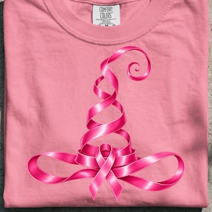 Könnte beinhalten: Ein hellrosa T-Shirt mit einer Grafik eines Hexenhutes, der aus einem durchgehenden rosa Band geformt ist. Das Etikett am Halsausschnitt zeigt COMFORT COLORS, HEAVYWEIGHT GARMENT DYE, RING SPUN, 100% COTTON, MADE IN HONDURAS, M-M-M.