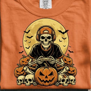 Op de afbeelding: Een oranje t-shirt met een afbeelding van een skelet dat een honkbalpet en een koptelefoon draagt terwijl het een videogame speelt. Het skelet is omringd door pompoenen, schedels, vleermuizen en een grote volle maan op de achtergrond.